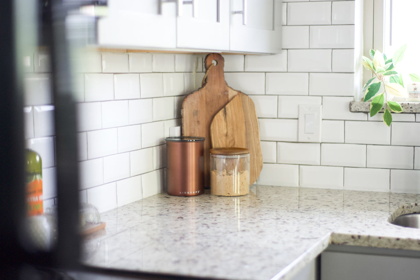 A kitchen countertop with a white subway tile backsplash, featuring two wooden cutting boards leaning against the wall, a copper canister, and a clear jar with a wooden lid. A blurred plant is visible on the windowsill to the right.