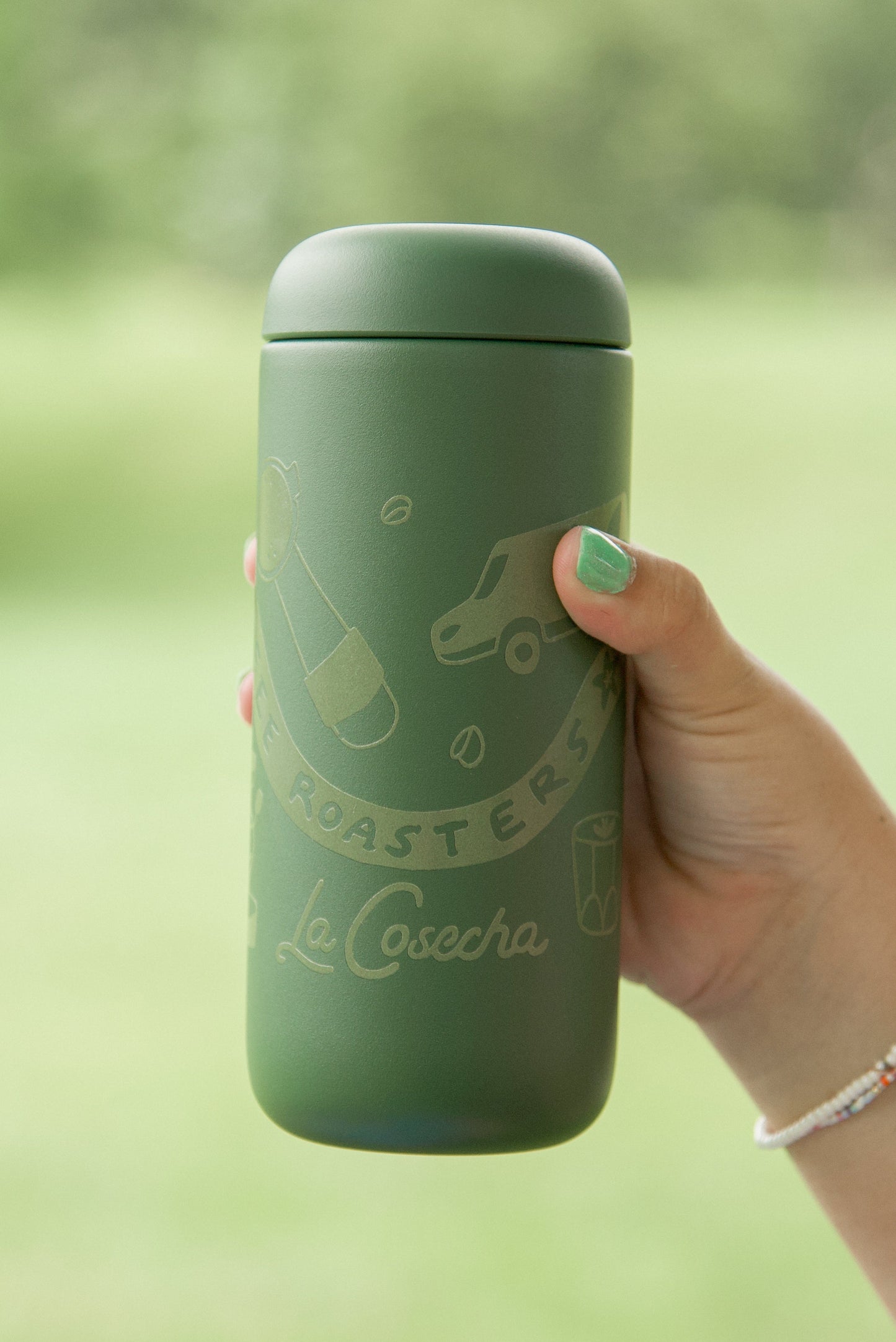 Hand holding a green thermos with 'La Cosecha' branding against a blurred natural background