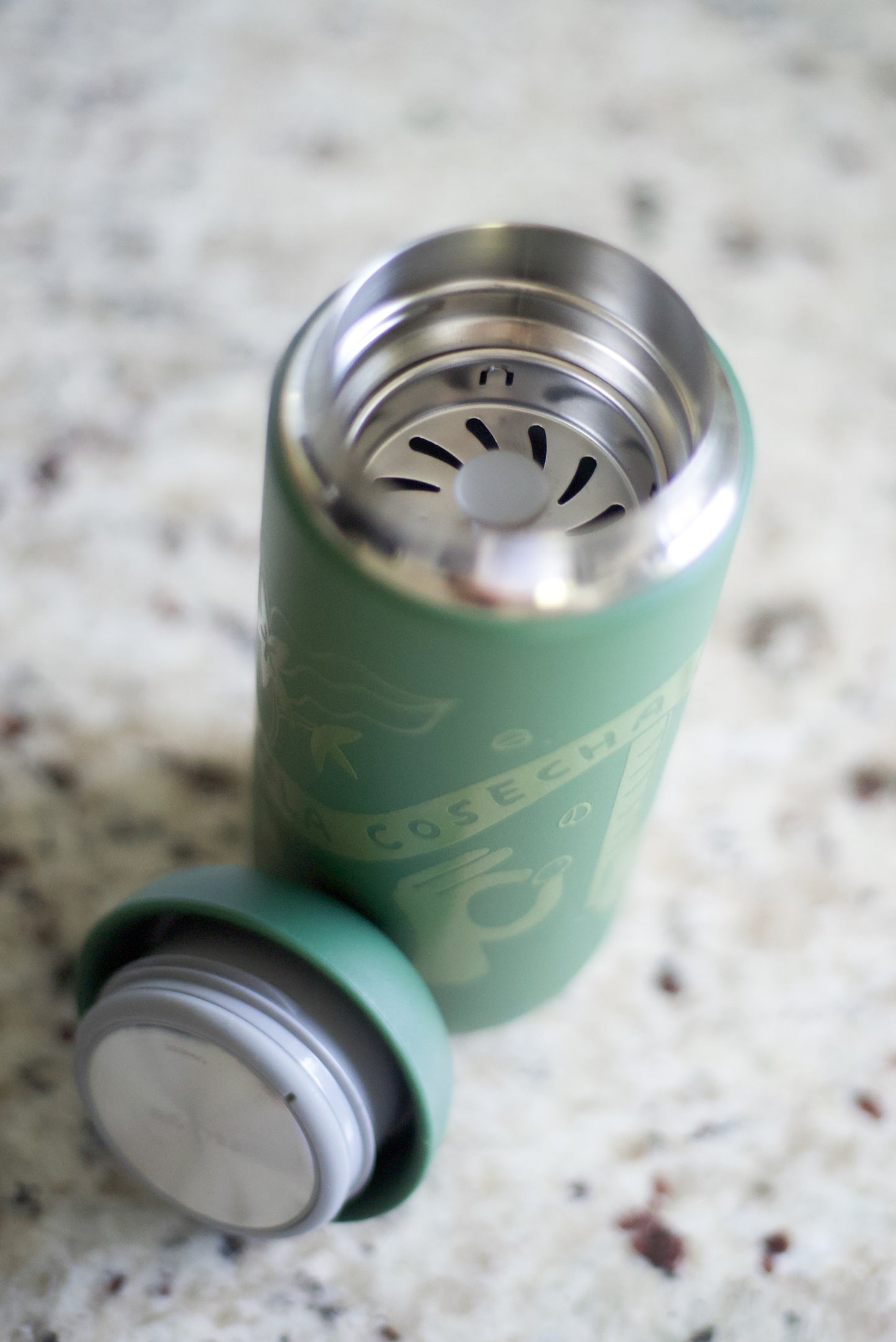 A green reusable travel tumbler with a matching lid placed beside it on a speckled stone countertop. The tumbler is open, showing a stainless steel interior with a strainer insert, and the lid has a silver metal rim.