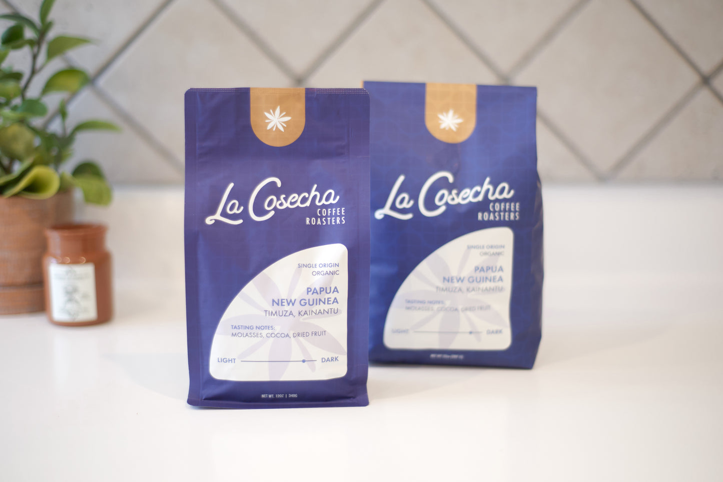 Two blue coffee bags labeled 'La Cosecha' on a white surface with a plant in the background.
