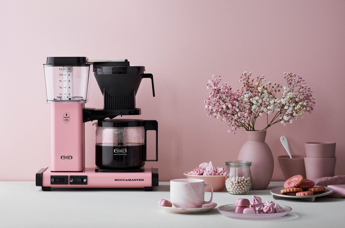 Pink and black coffee maker on a table with a pink wall and decorative items.