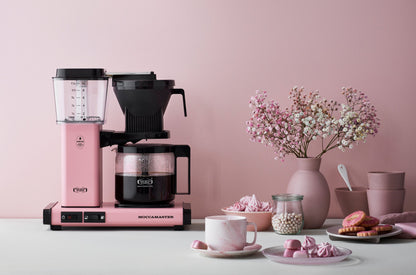 Pink and black coffee maker on a table with a pink wall and decorative items.