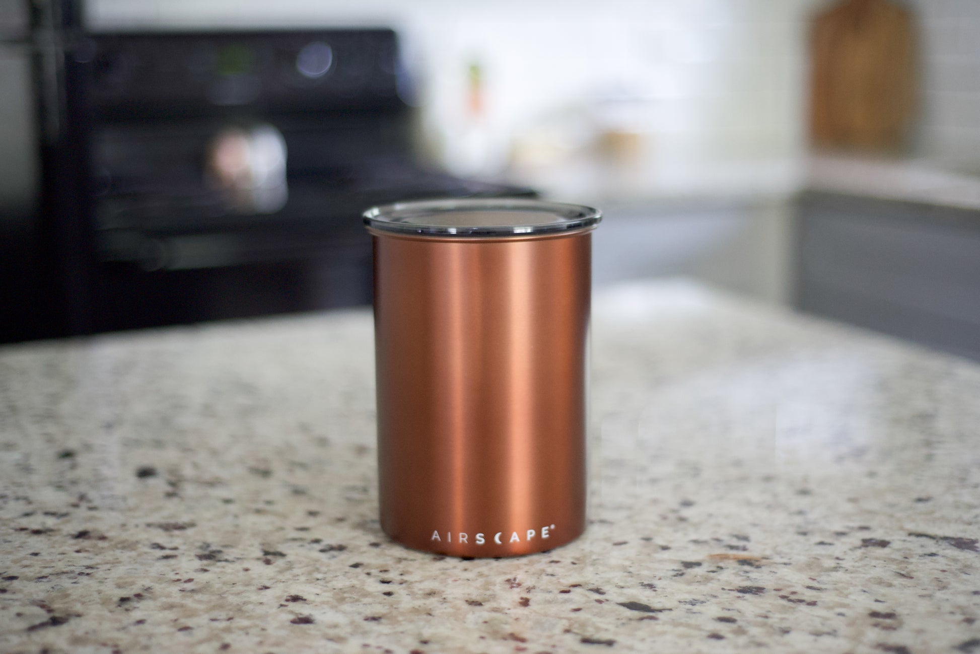 A copper-colored Airscape insulated tumbler with a clear lid, standing on a speckled granite kitchen countertop with a blurred kitchen background.