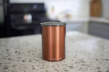 A copper-colored Airscape insulated tumbler with a clear lid, standing on a speckled granite kitchen countertop with a blurred kitchen background.