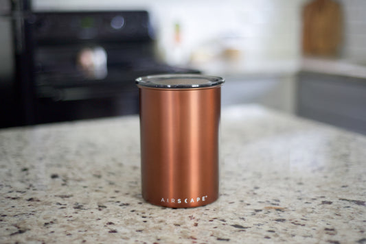 A copper-colored Airscape insulated tumbler with a clear lid, standing on a speckled granite kitchen countertop with a blurred kitchen background.