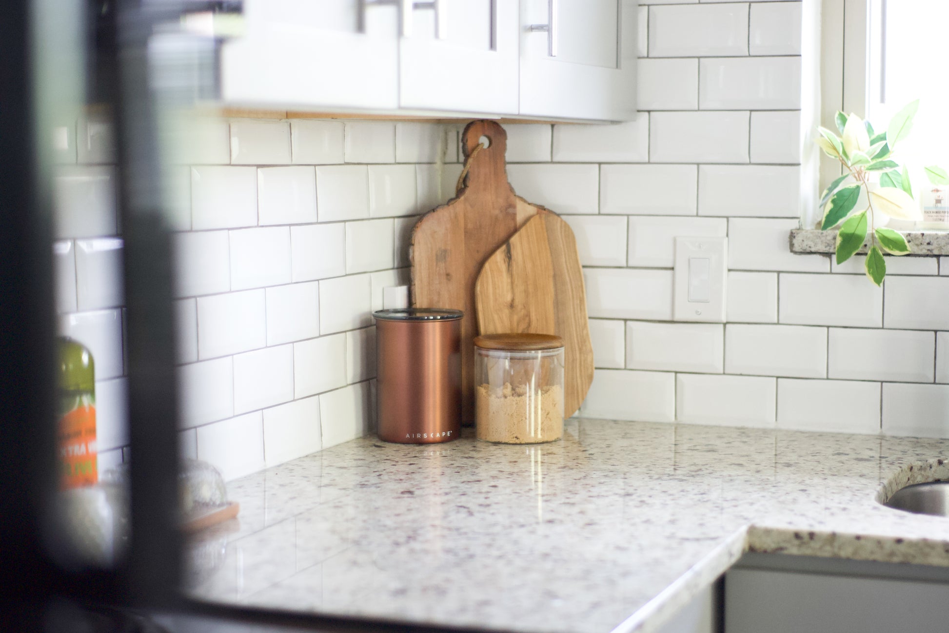 A kitchen countertop with a white subway tile backsplash, featuring two wooden cutting boards leaning against the wall, a copper canister, and a clear jar with a wooden lid. A blurred plant is visible on the windowsill to the right.