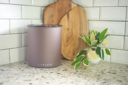 A matte dusty rose cylindrical container with a silver lid sits on a speckled granite countertop in a kitchen setting, with a white subway tile backsplash, wooden cutting boards, and a small potted variegated plant in the background.