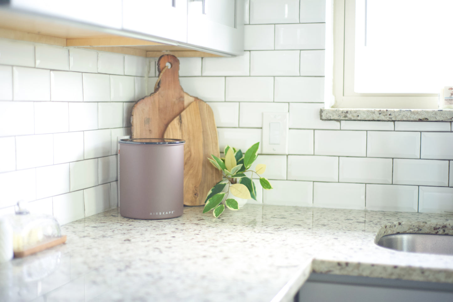 A matte taupe metal canister with the brand name 'Airscape' visible on the front, styled on a speckled granite kitchen counter next to a wooden cutting board and a small green plant.