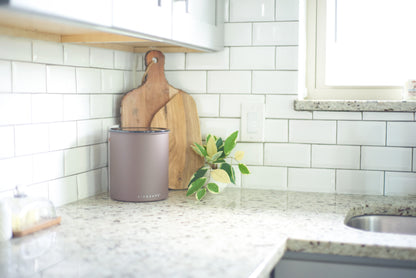 A matte taupe metal canister with the brand name 'Airscape' visible on the front, styled on a speckled granite kitchen counter next to a wooden cutting board and a small green plant.