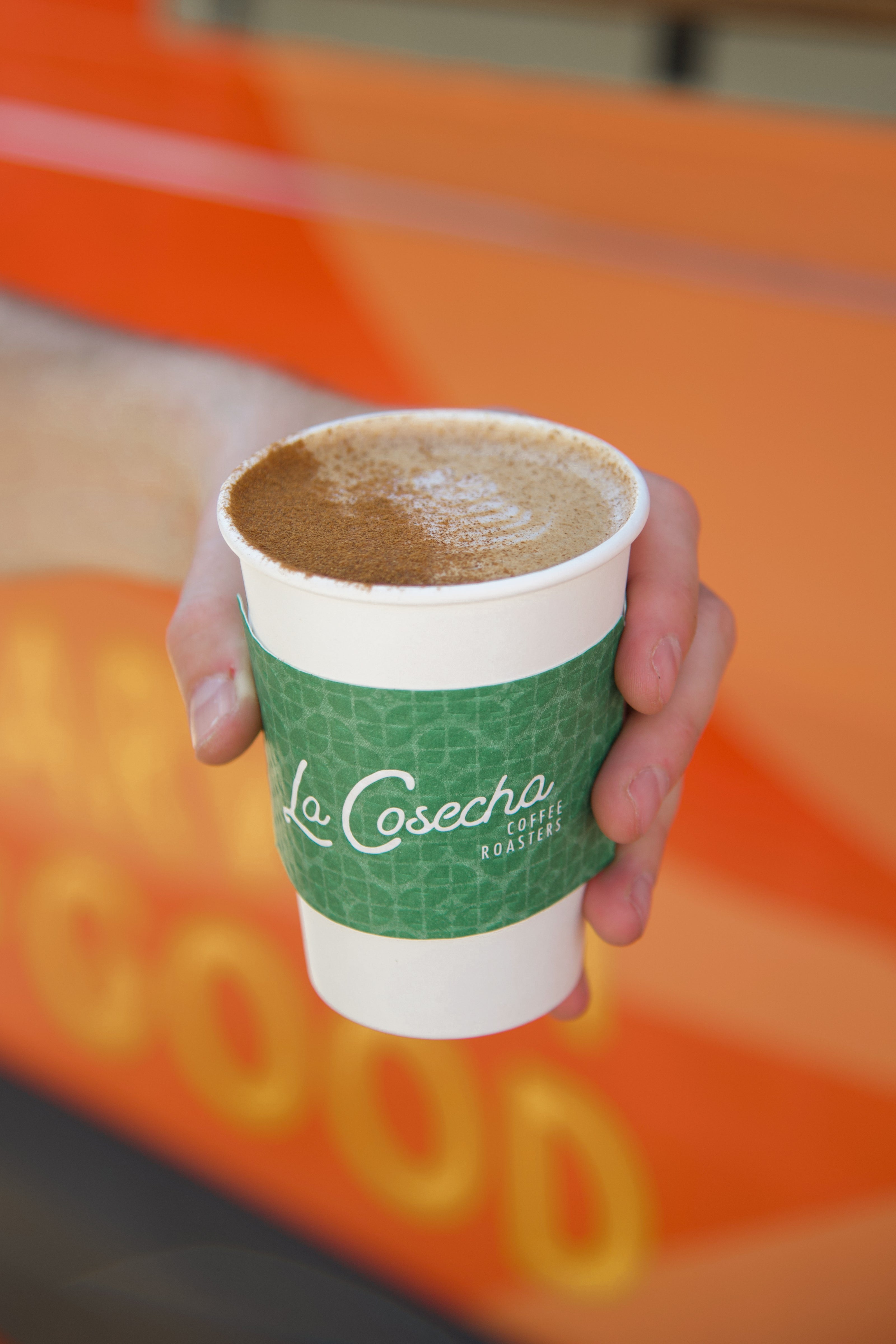 Hand holding a green and white coffee cup with 'La Cosecha' branding against an orange background.