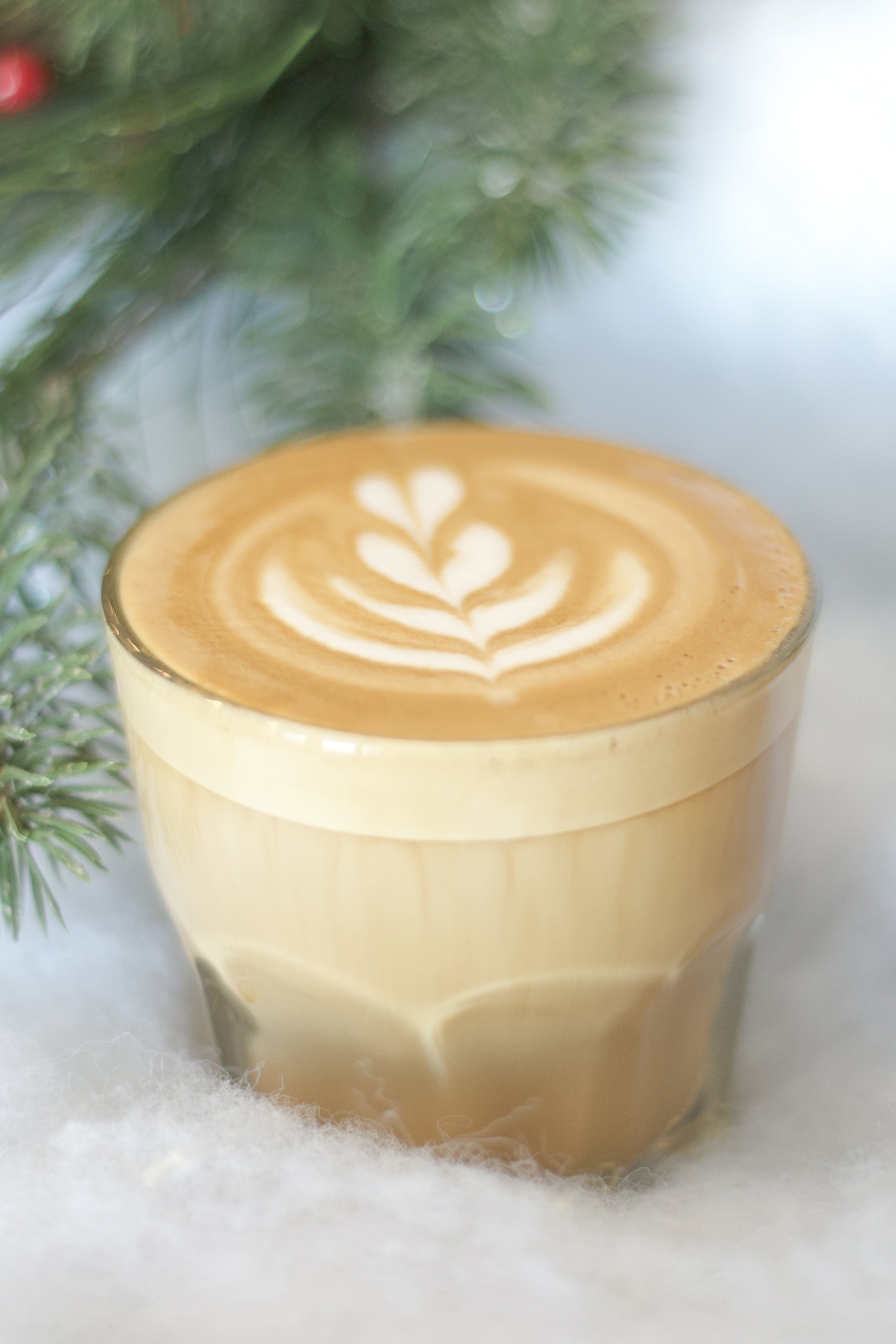 Glass of latte with decorative foam art on a snowy background with pine branches and pinecones.