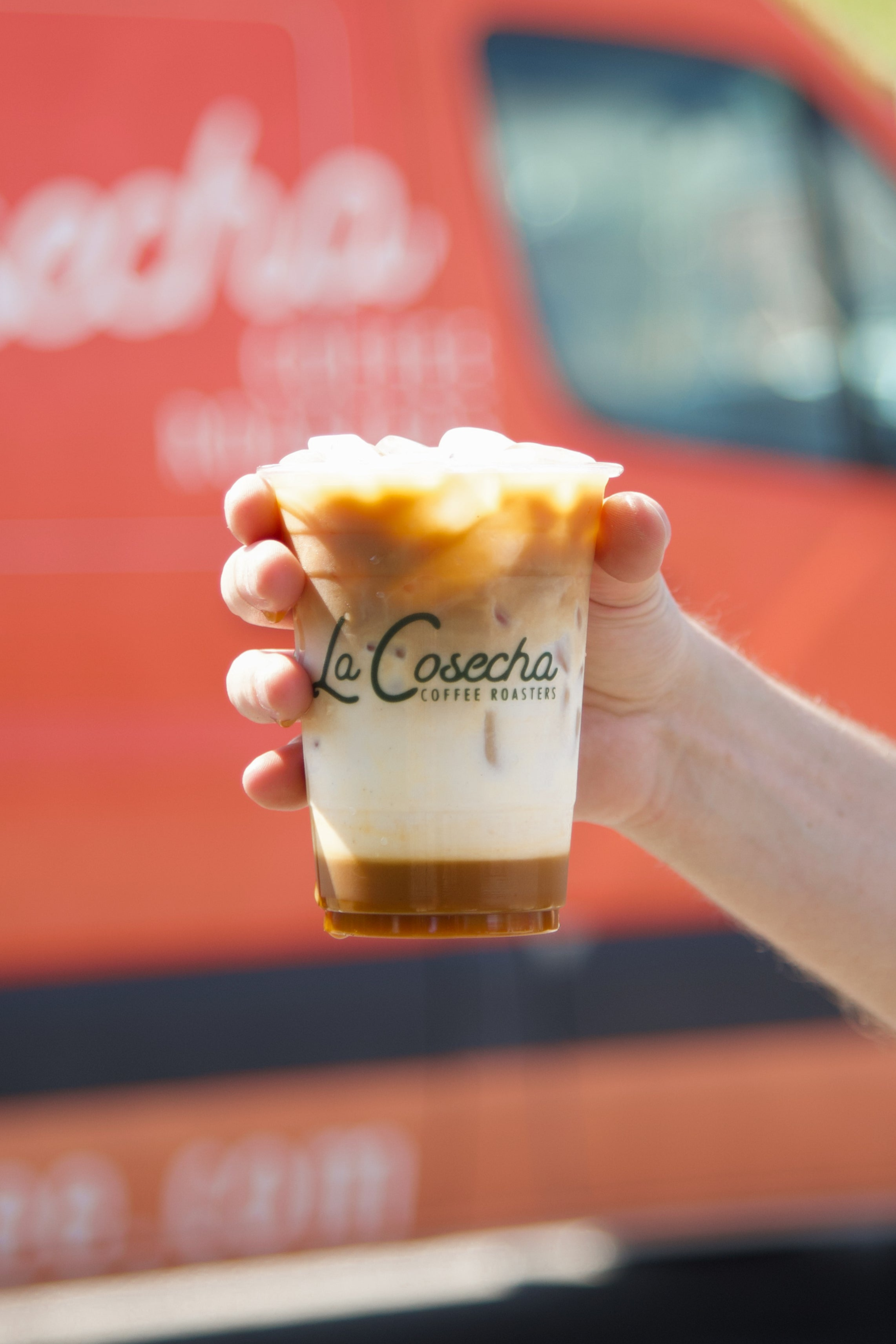 Hand holding a cup of coffee with 'La Cosecha' branding in front of a red vehicle.