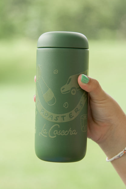 Hand holding a green thermos with 'La Cosecha' branding against a blurred natural background