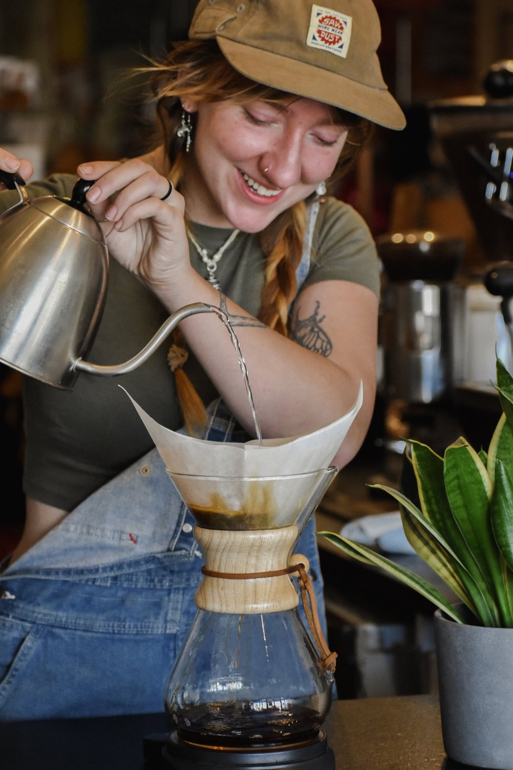 Person pouring coffee from a kettle into a Chemex in a coffee shop setting.