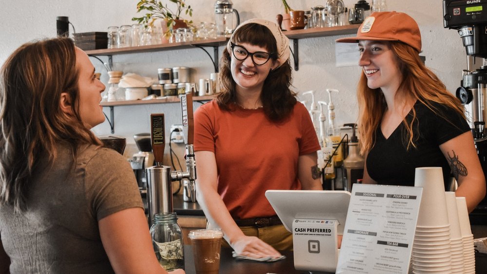 Three women in a coffee shop with a laptop and menu.