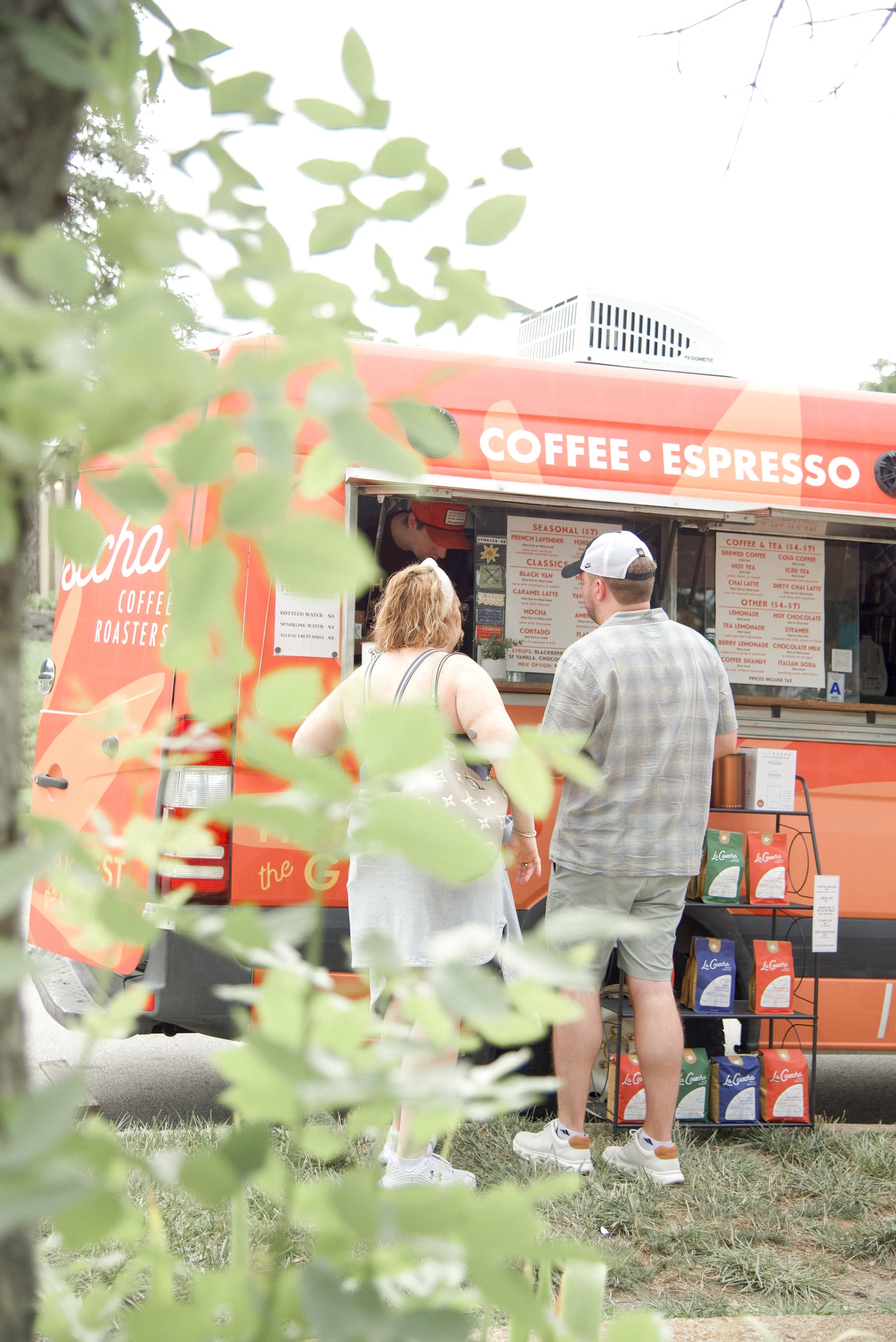 Two people standing in front of a coffee and espresso food truck with greenery in the foreground.