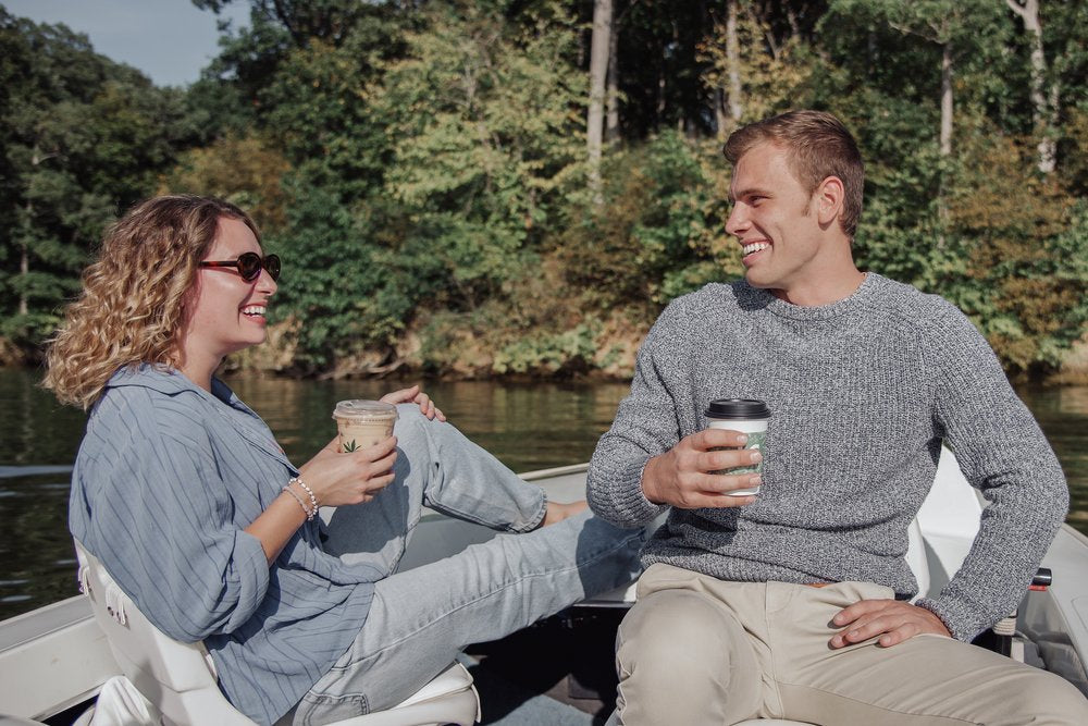 Two people sitting on a boat by a river, holding coffee cups and smiling.
