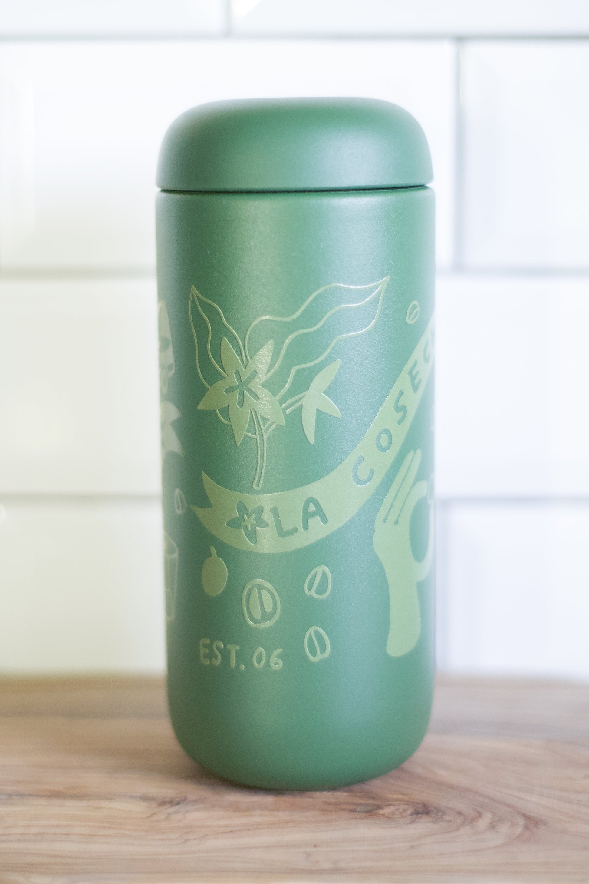 A matte sage green insulated tumbler with a matching lid, featuring light sage green etched illustrations of coffee beans, a leaf, and a hand with the text 'LA COFFEE' and 'EST. 06', standing on a wooden surface against a white tiled background.