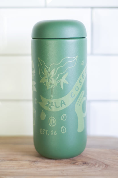 A matte sage green insulated tumbler with a matching lid, featuring light sage green etched illustrations of coffee beans, a leaf, and a hand with the text 'LA COFFEE' and 'EST. 06', standing on a wooden surface against a white tiled background.