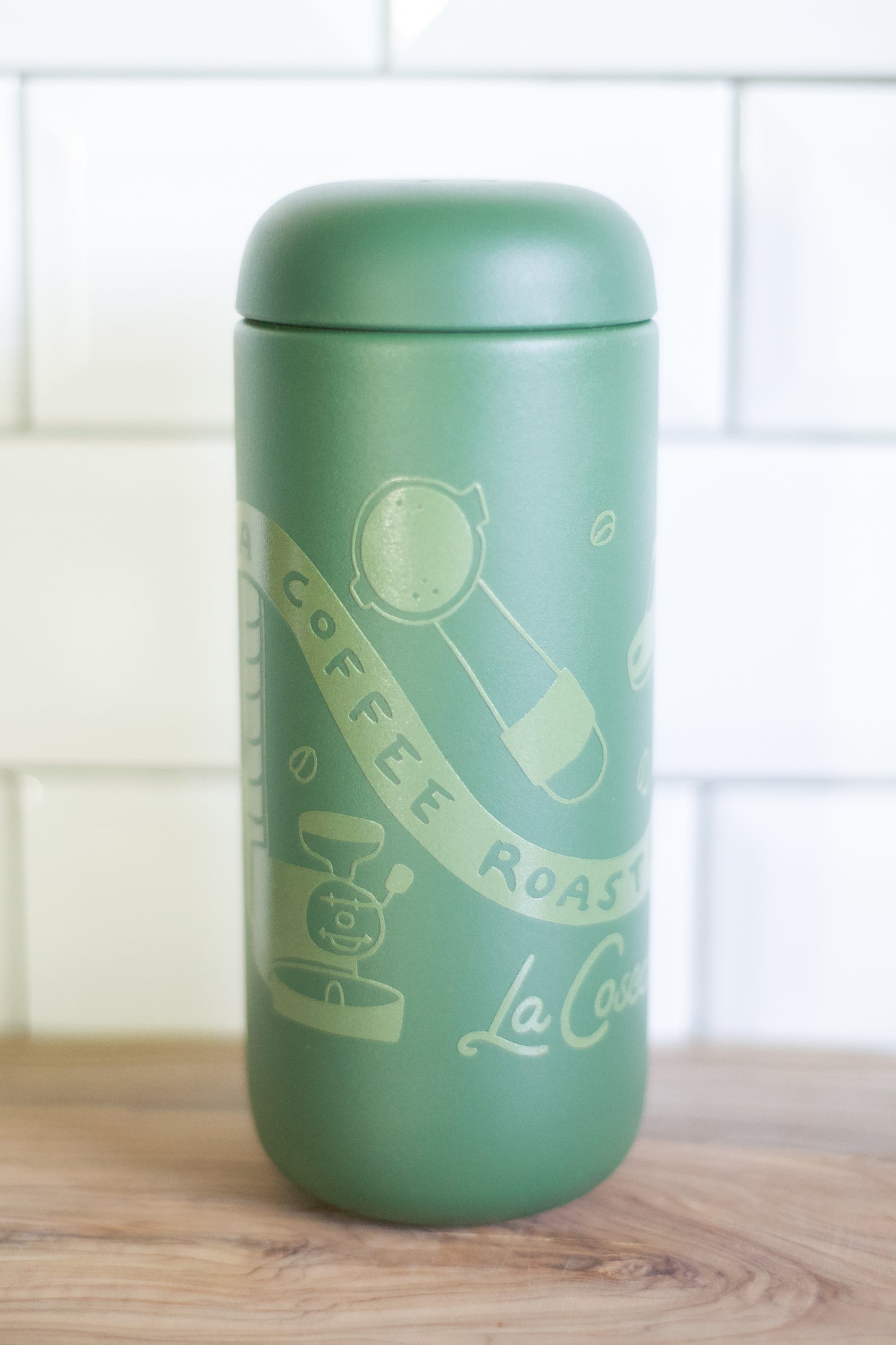 A sage green insulated coffee cup with a matching lid, featuring light green illustrations of a coffee bean, a coffee grinder, and the text 'COFFEE ROASTING La Cosecha on the front, set on a wooden surface against a white tiled background.