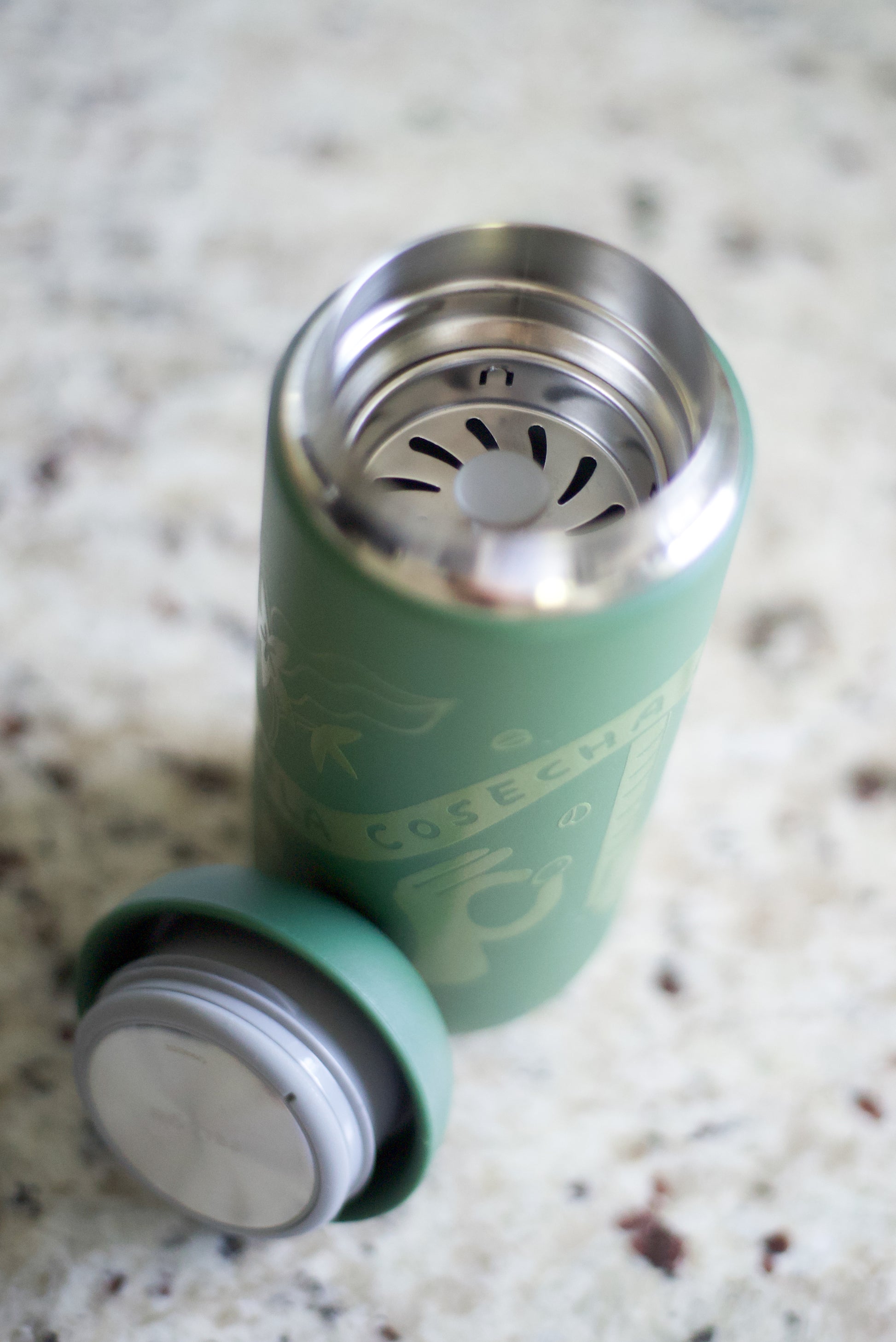 A green reusable travel tumbler with a matching lid placed beside it on a speckled stone countertop. The tumbler is open, showing a stainless steel interior with a strainer insert, and the lid has a silver metal rim.