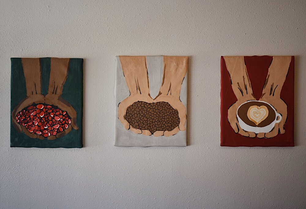 Three paintings of hands holding coffee beans and a cup on a white wall.