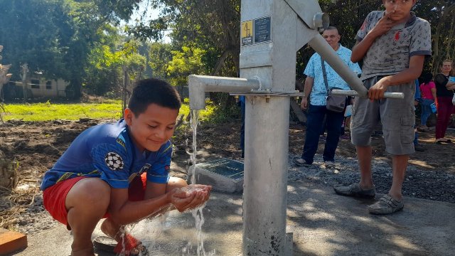 Child washing hands at a water pump with people in the background. Living Water International