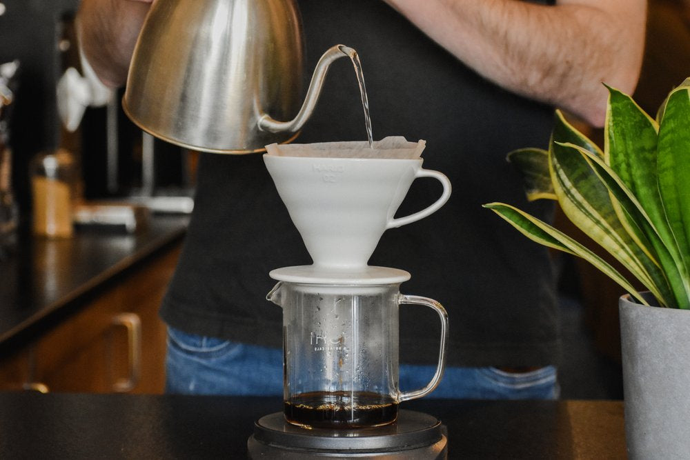 Person making coffee using a pour-over method with a white coffee filter and glass carafe.