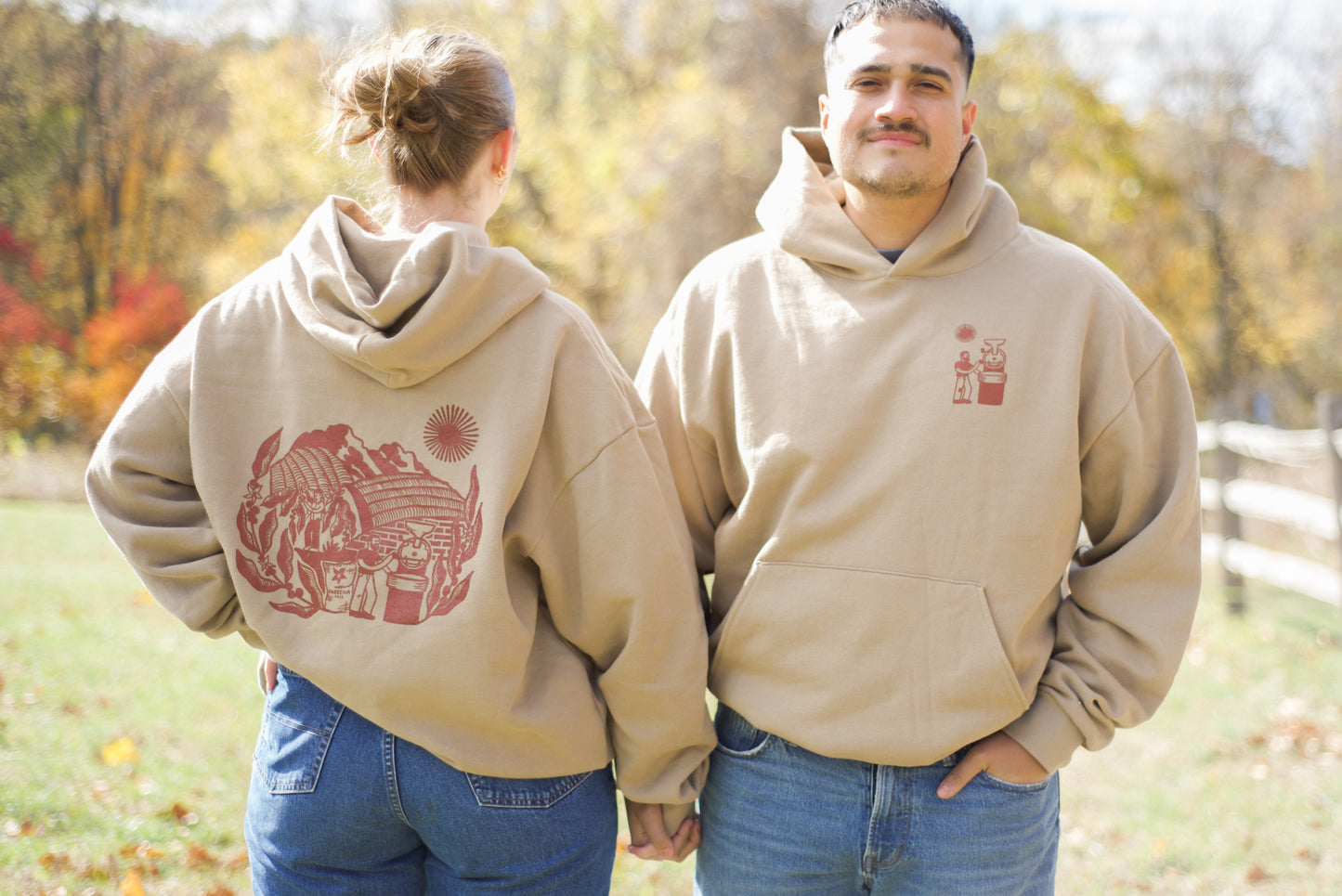 Two people wearing beige hoodies with red graphics in a park setting.