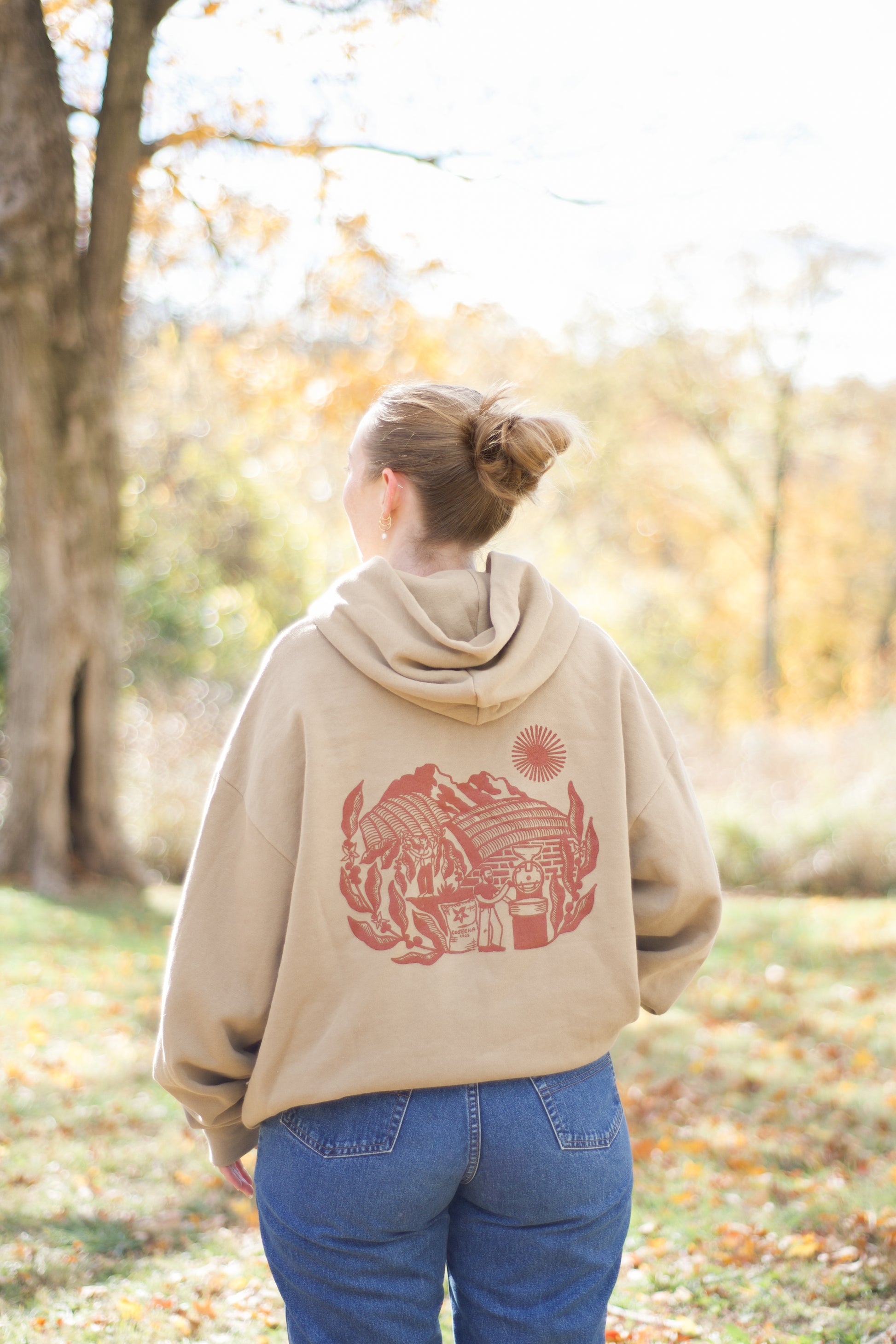 Person wearing a beige hoodie with a red graphic design in an outdoor setting with trees and grass.