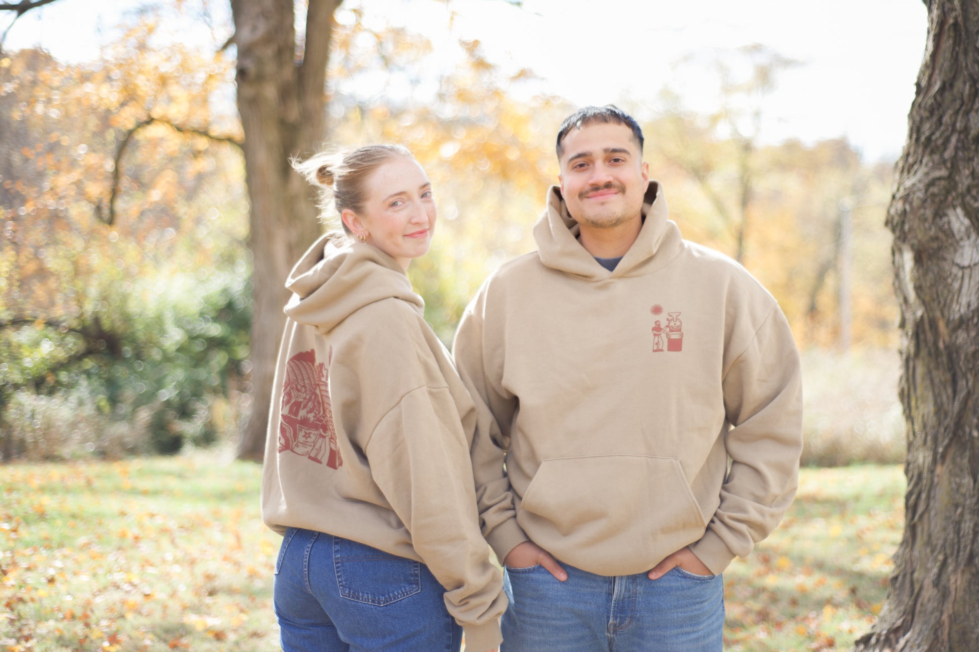 Two people wearing matching beige hoodies with a logo in an outdoor setting with trees and grass.