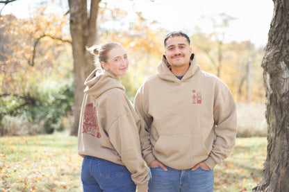 Two people wearing matching beige hoodies with a logo in an outdoor setting with trees and grass.