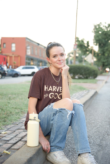 Person wearing a brown t-shirt with Harvest the Good text, sitting on a curb outdoors.