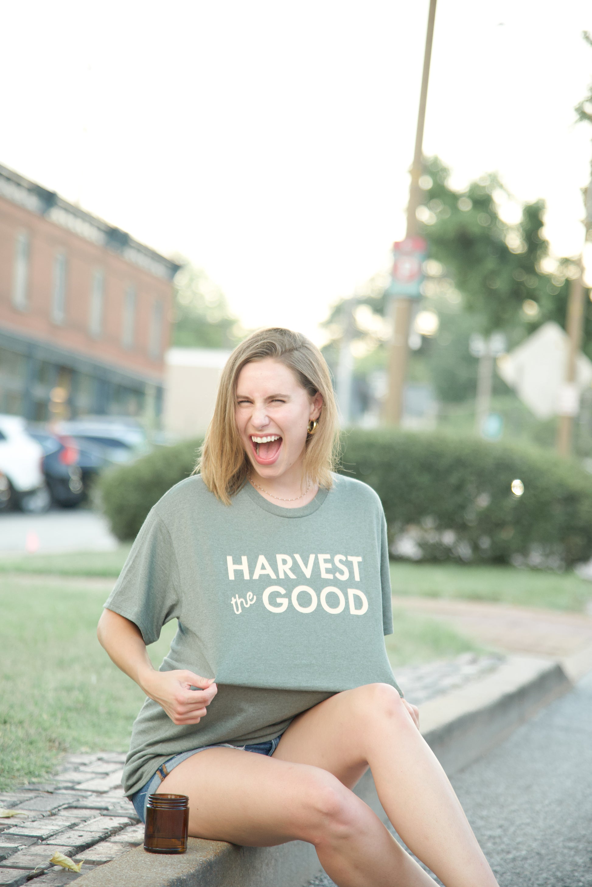Woman wearing a 'Harvest the Good' t-shirt sitting on a curb outdoors.