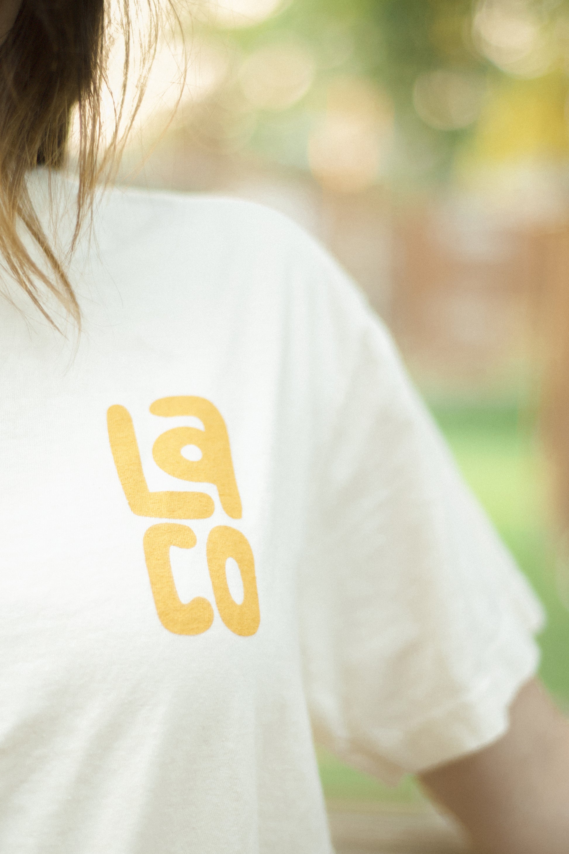 A close-up shot of a person wearing a white t-shirt with a yellow 'LA CO' graphic printed on the back, set against a blurred outdoor background.
