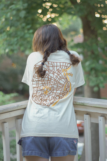 A person viewed from behind wearing a cream-colored t-shirt with a large brown and yellow street map graphic on the back, paired with denim shorts, standing on a wooden deck with a lush green outdoor background.