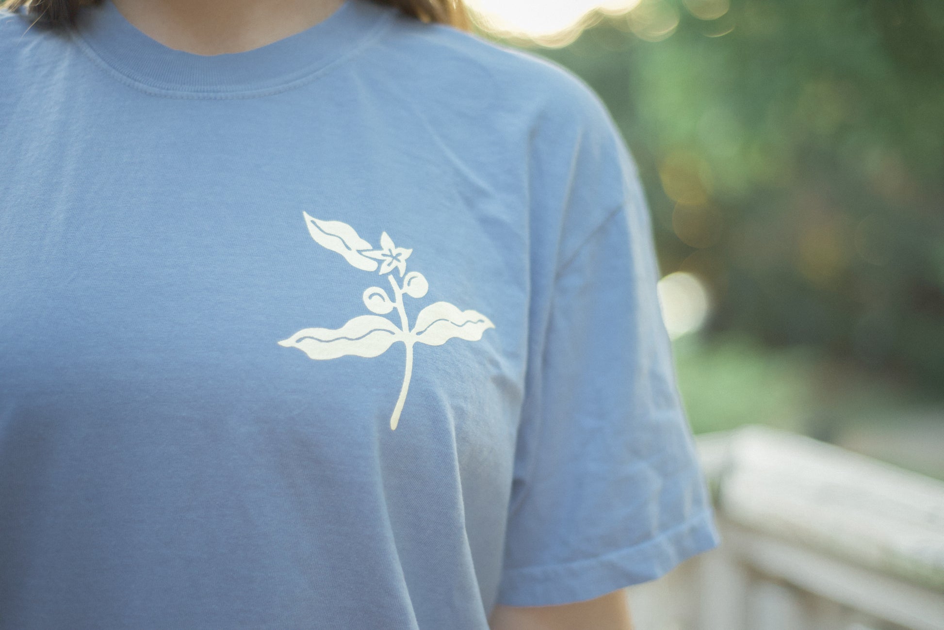 A close-up of a person wearing a light blue crewneck t-shirt with a white coffee plant graphic on the chest, set against a blurred outdoor background.