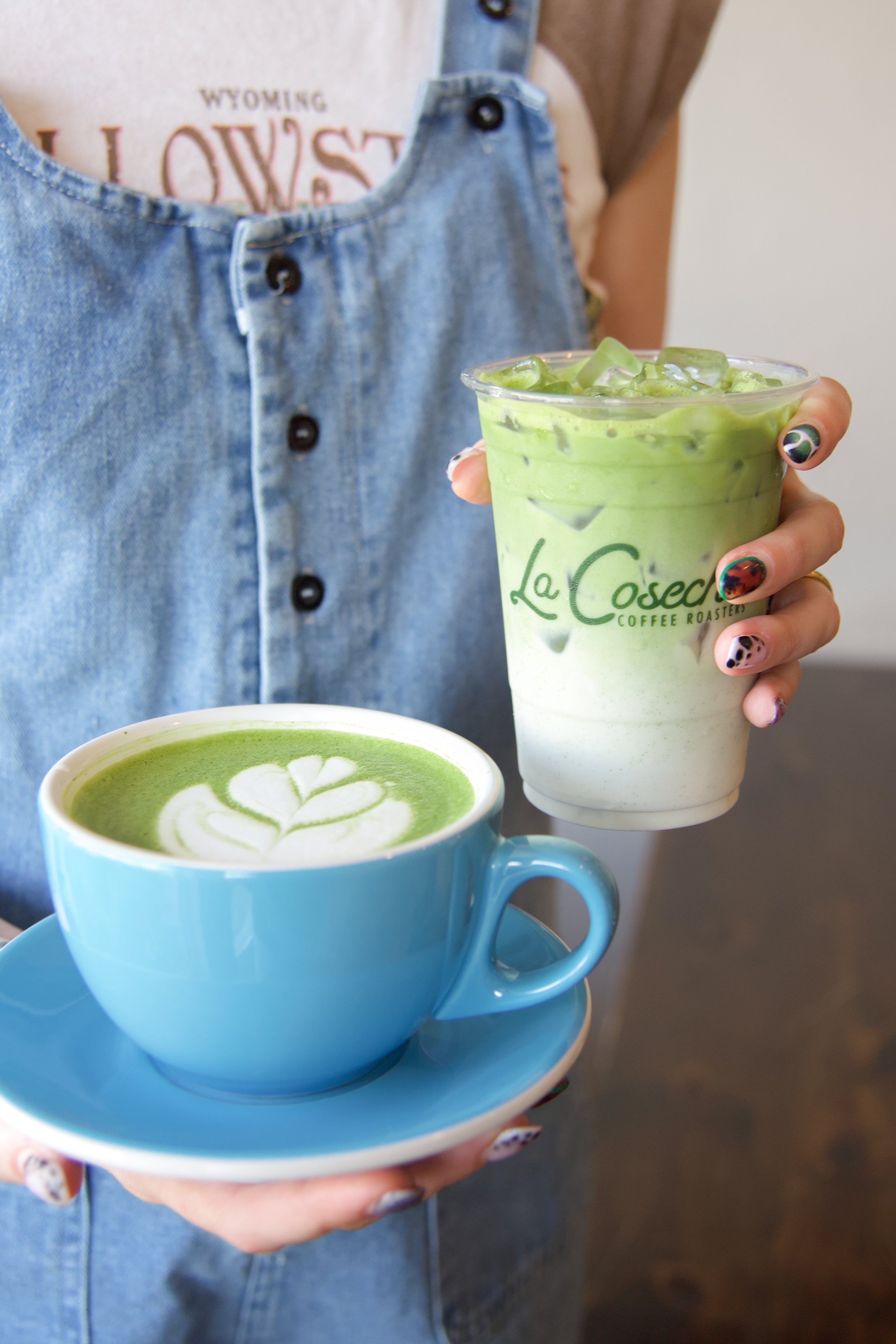 Person holding a blue cup and a clear plastic cup with green matcha drink, wearing a denim overalls.