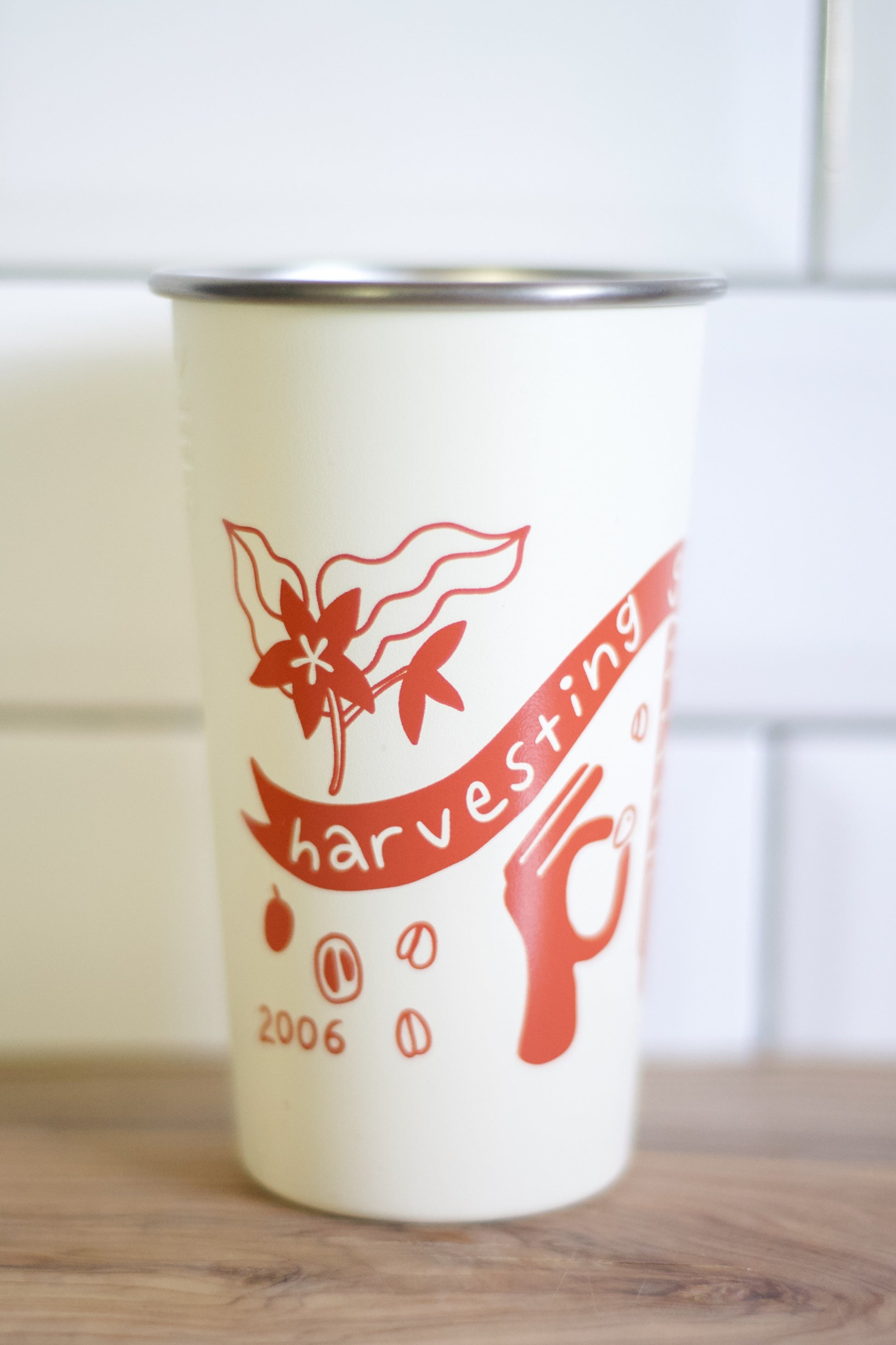 An off-white insulated coffee cup with a stainless steel rim, featuring a red graphic of coffee cherries, beans, and the word 'harvesting' on a banner, set against a tiled background.