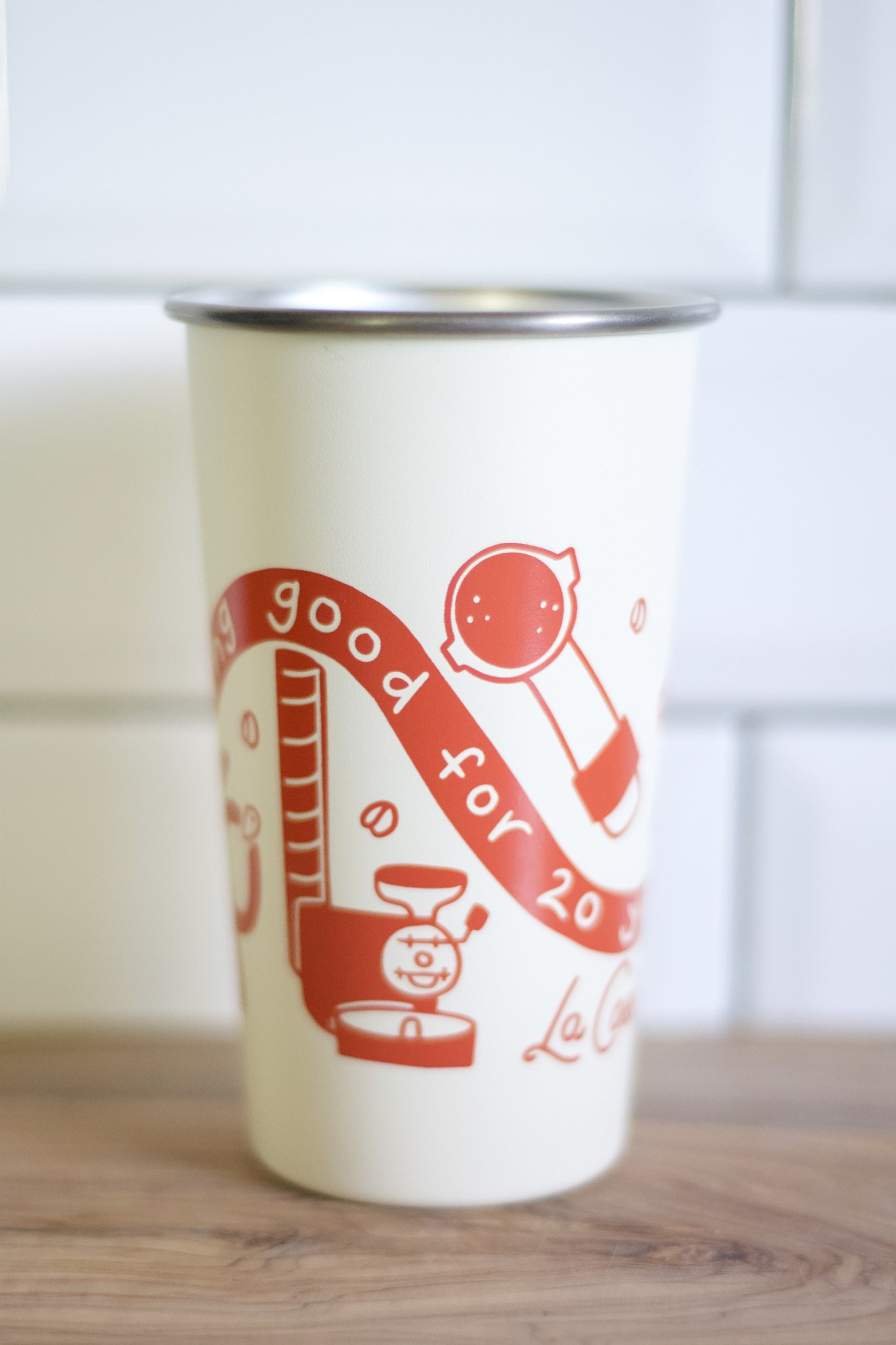 An off-white insulated travel tumbler with a stainless steel rim, featuring a red graphic illustration of a coffee grinder and a banner that reads 'good for 20'.