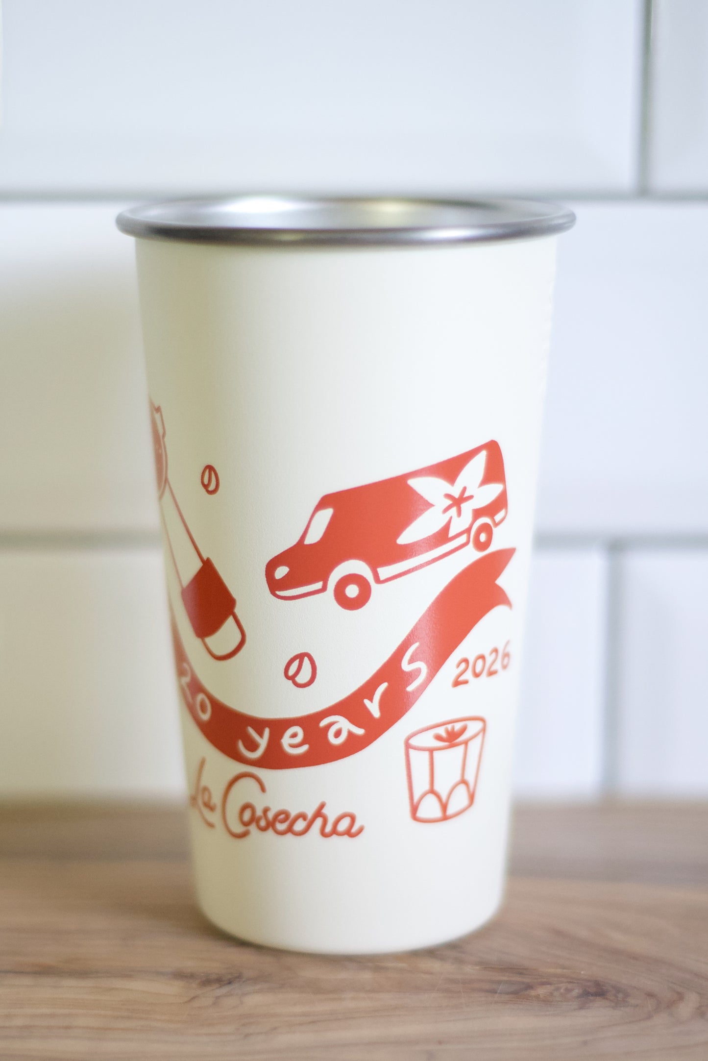 An off-white insulated travel tumbler with a stainless steel rim, featuring a red graphic print of a van, ribbon, gift box, and the text '20 years 2026' and 'La Cosecha'.