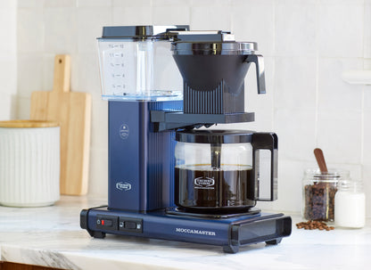 Blue Moccamaster coffee maker on a kitchen counter with a white tiled wall background.