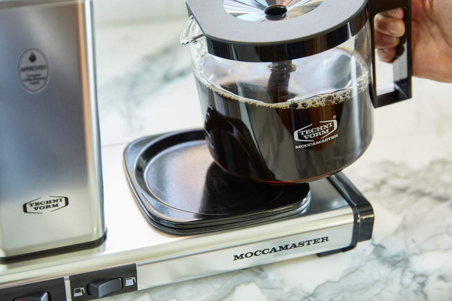 Moccamaster coffee maker on a marble countertop with a person holding the carafe.