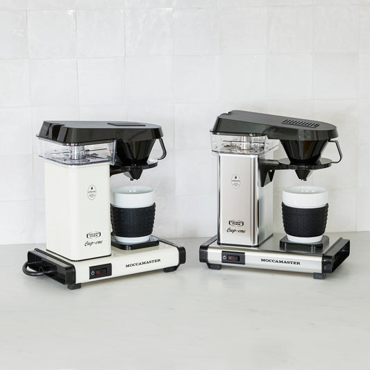 Two Moccamaster coffee makers on a white surface with a white tiled wall background.