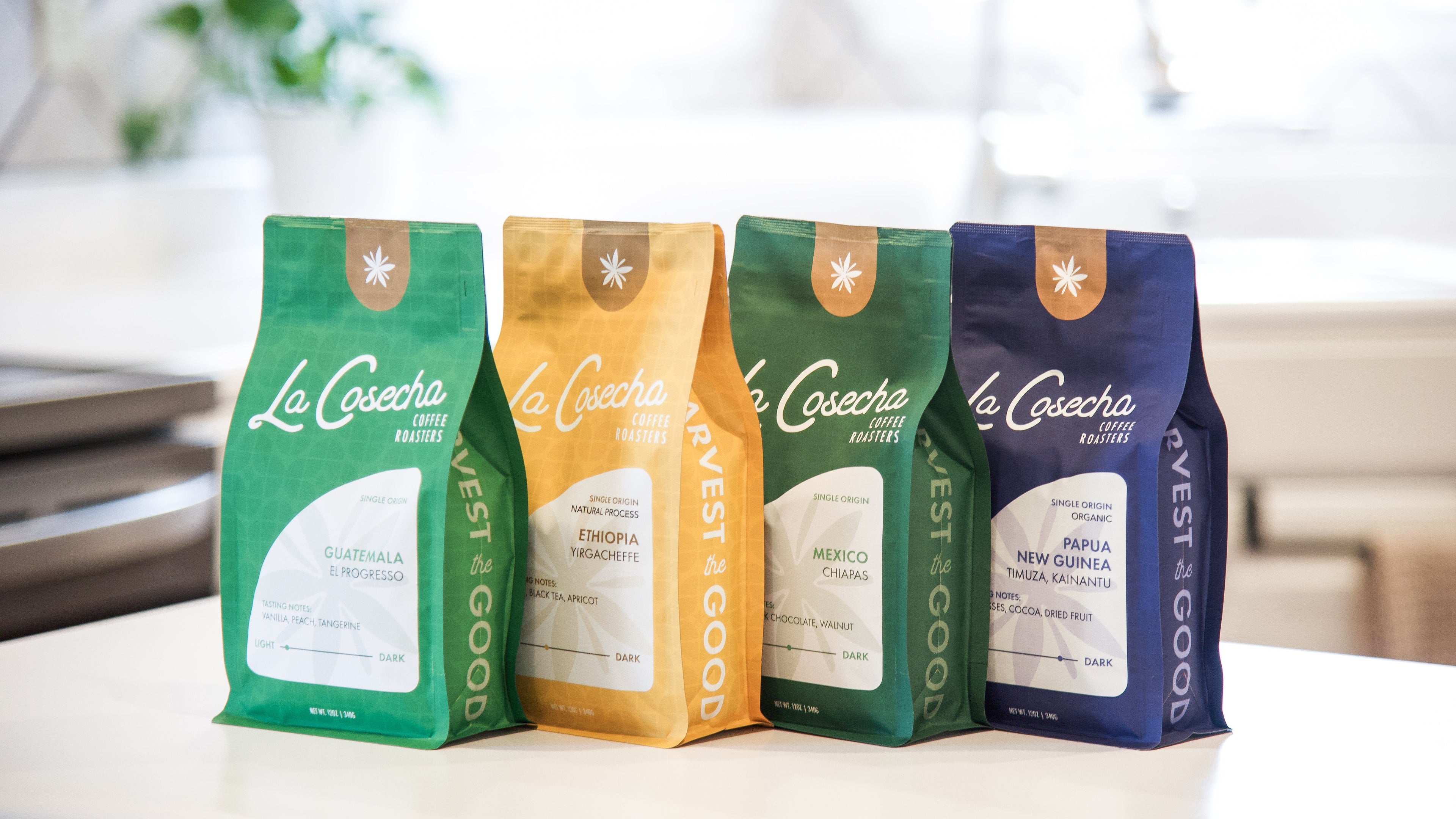 Four La Cosecha coffee bags in different colors on a white surface with a blurred background.