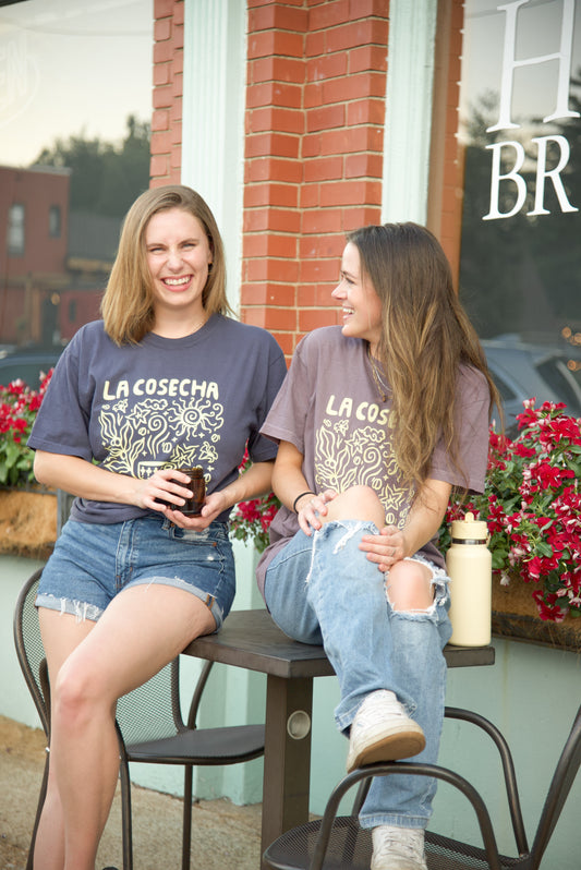 Two women wearing La Cosecha Coffee t-shirts sitting on a bench outside a building with flowers in the background.