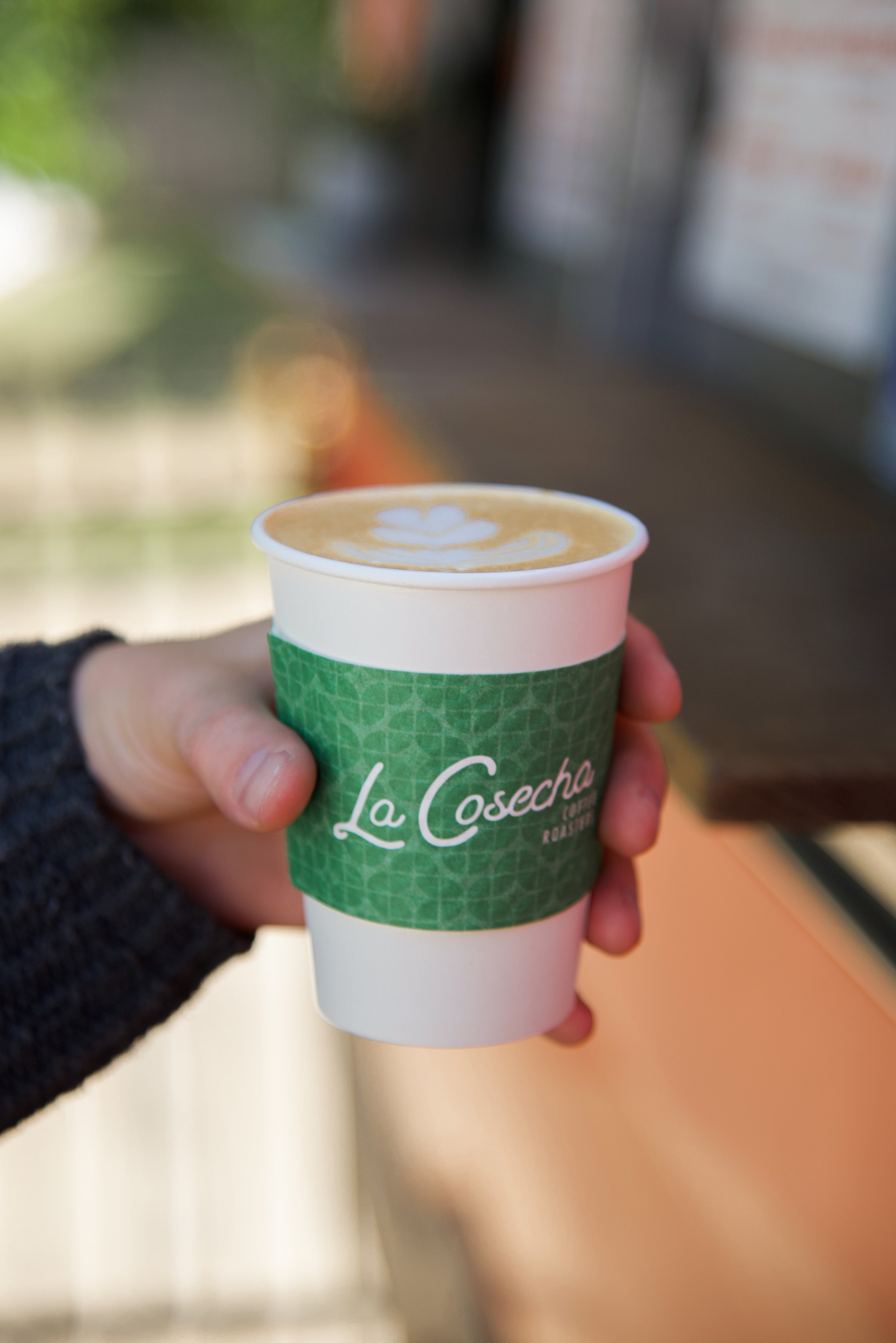 Hand holding a coffee cup with 'La Cosecha' branding outdoors.