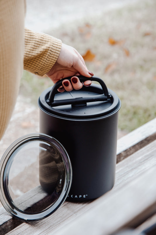 Person holding a black Airscape kilo with a transparent lid on a wooden surface.