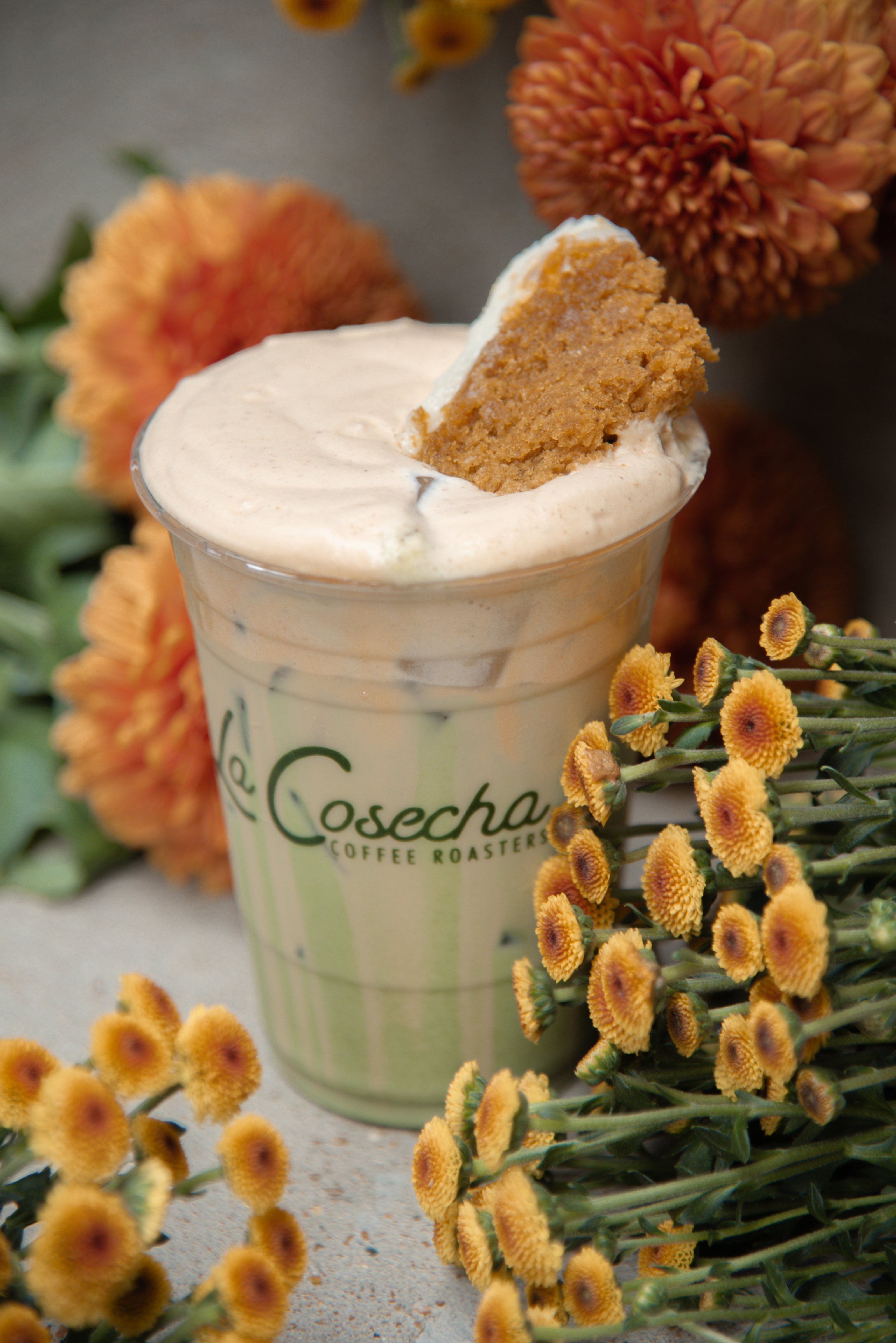 Green cup with 'La Cosecha' branding, topped with whipped cream and a cookie, surrounded by flowers.