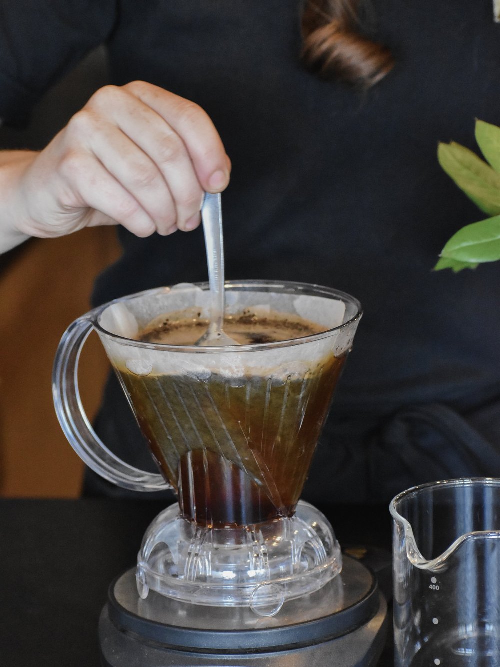 Person stirring a coffee filter with coffee grounds in a clear glass container.